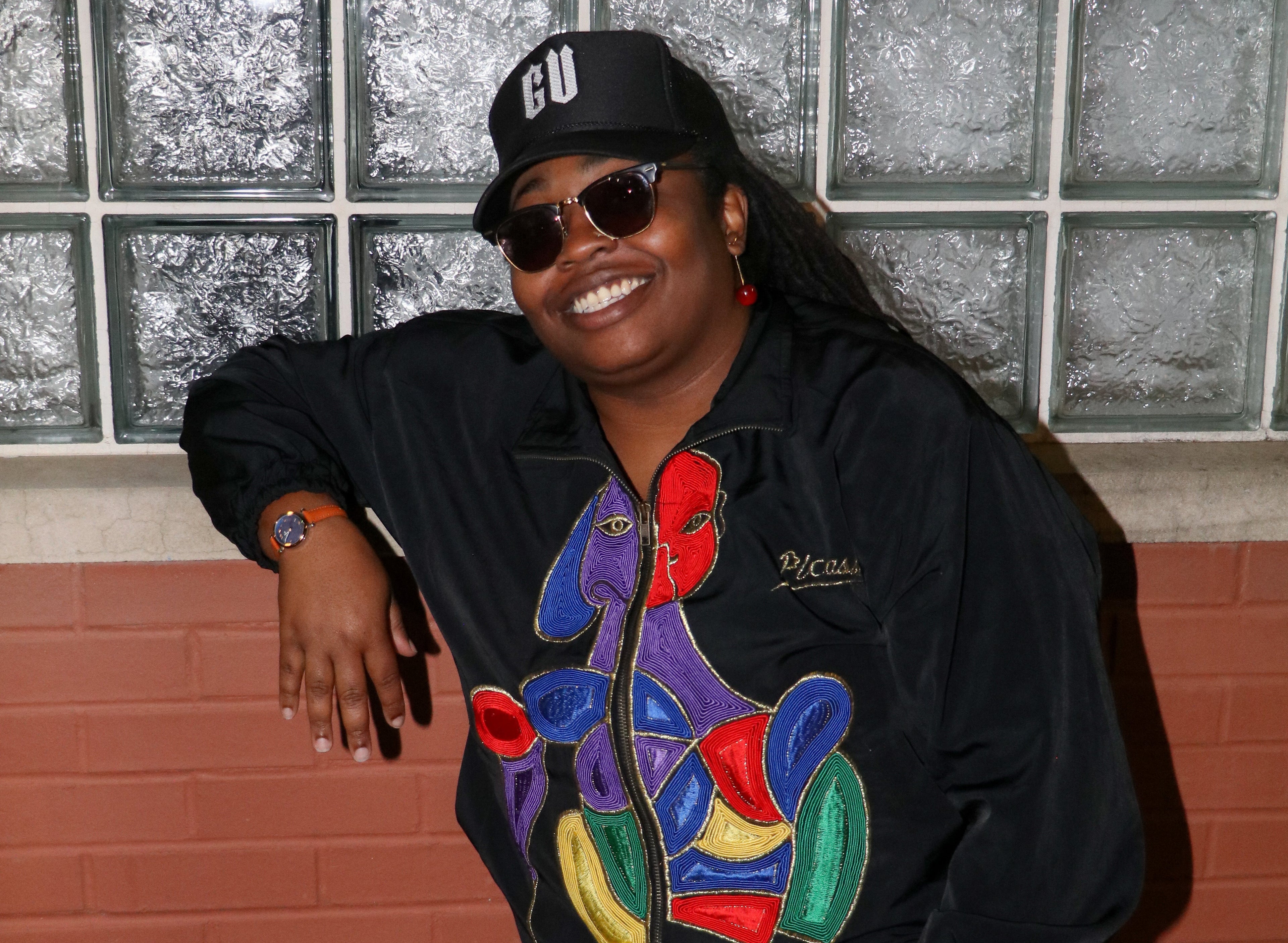 Person wearing a colorful jacket and red shorts standing against a glass block wall.