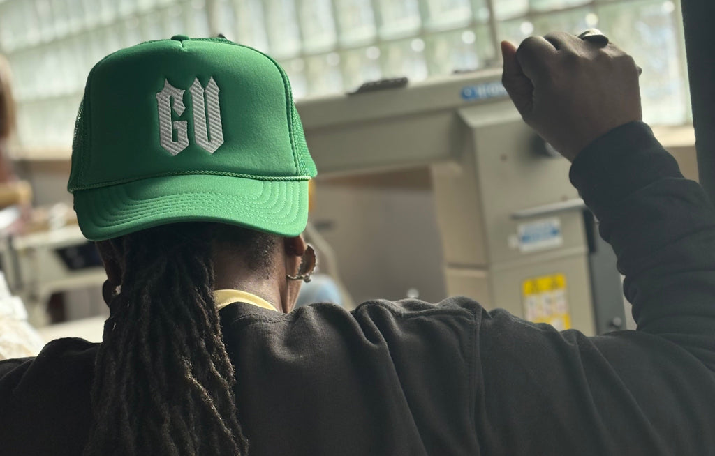Person wearing a green cap with 'GU' logo, sitting indoors with large windows in the background.